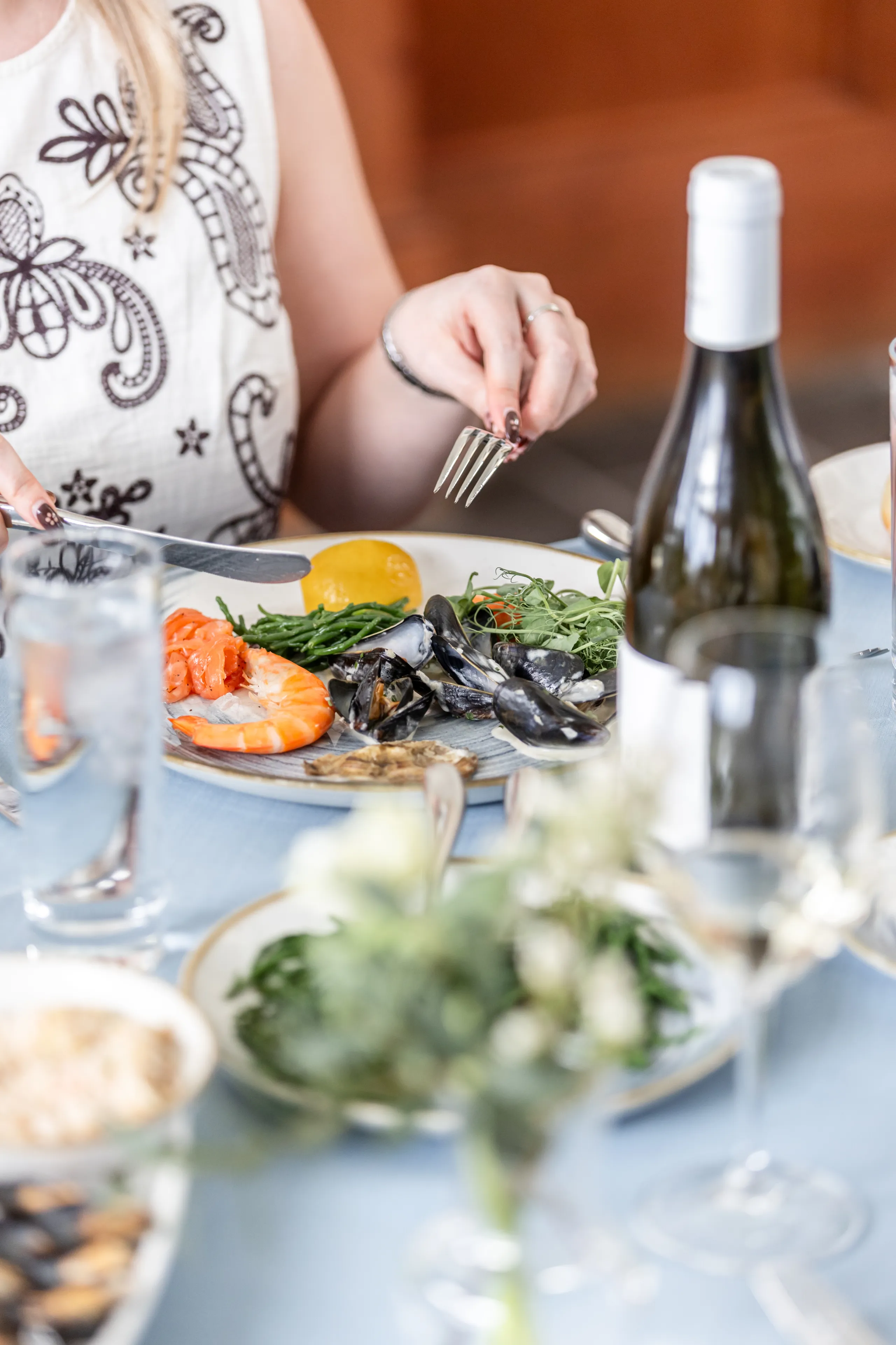 Woman dining on a seafood platter with prawns, mussels and greens at a restaurant table with a bottle of white wine.