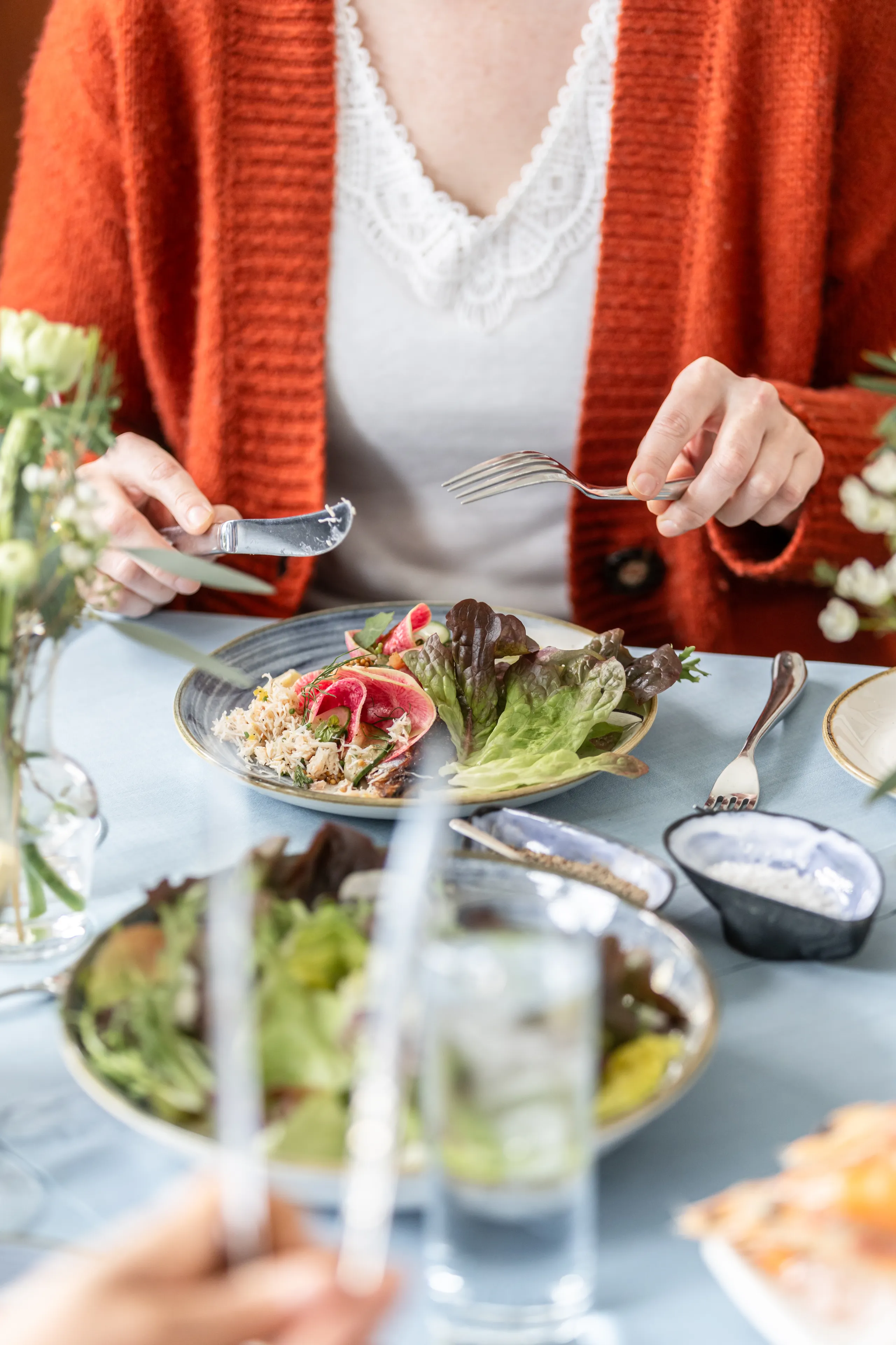Person in a rust-coloured cardigan holding a knife and fork over a plated salad at a restaurant table