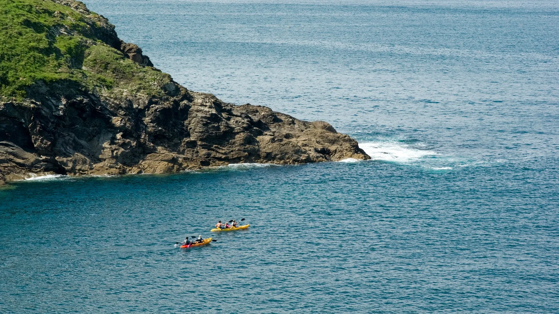 Two people kayaking on calm blue water beside a rocky, grass-covered coastline at Port Isaac, Cornwall.