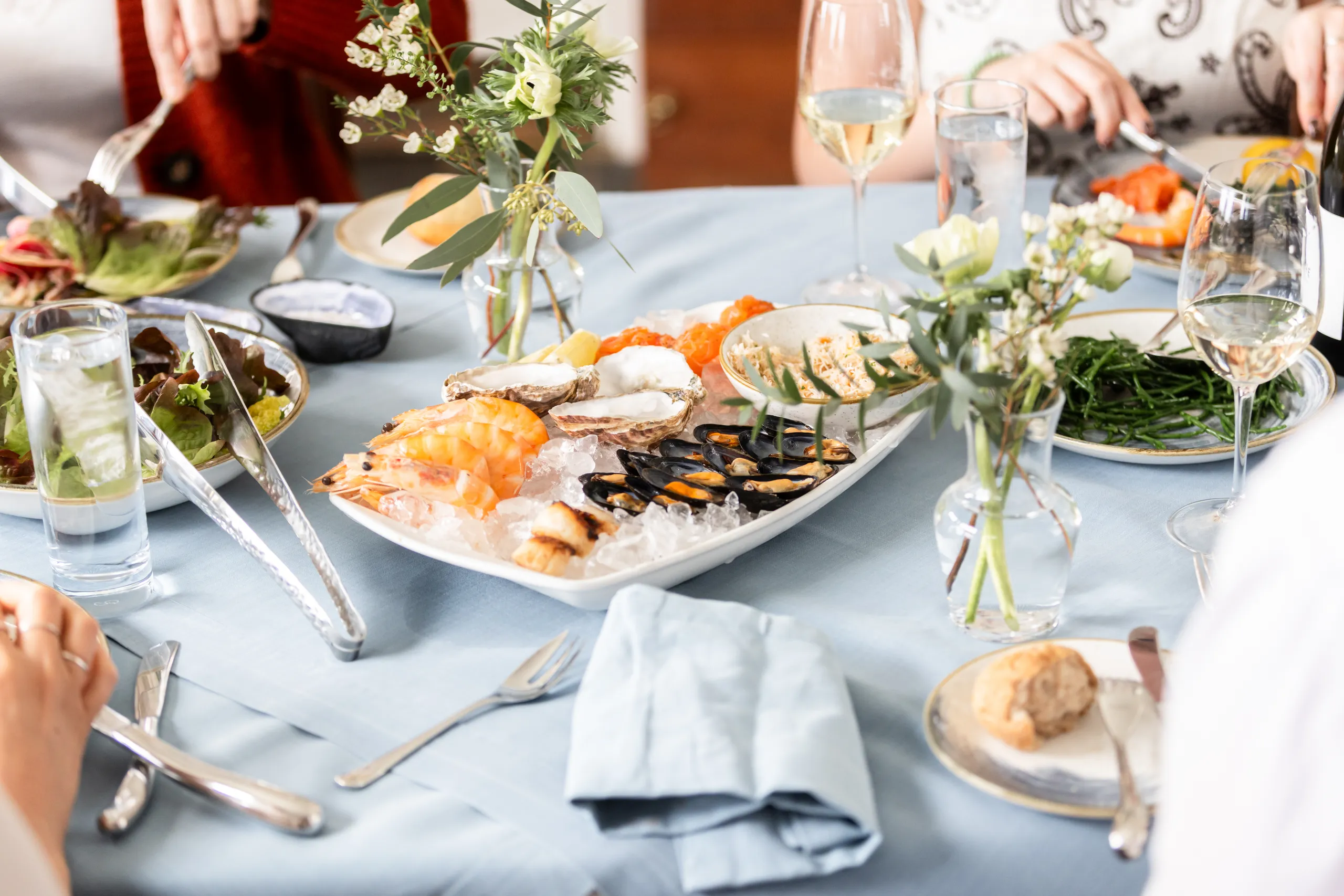 Elegant seafood dining table with prawns, oysters and mussels served on ice, surrounded by salads, wine glasses and small floral arrangements