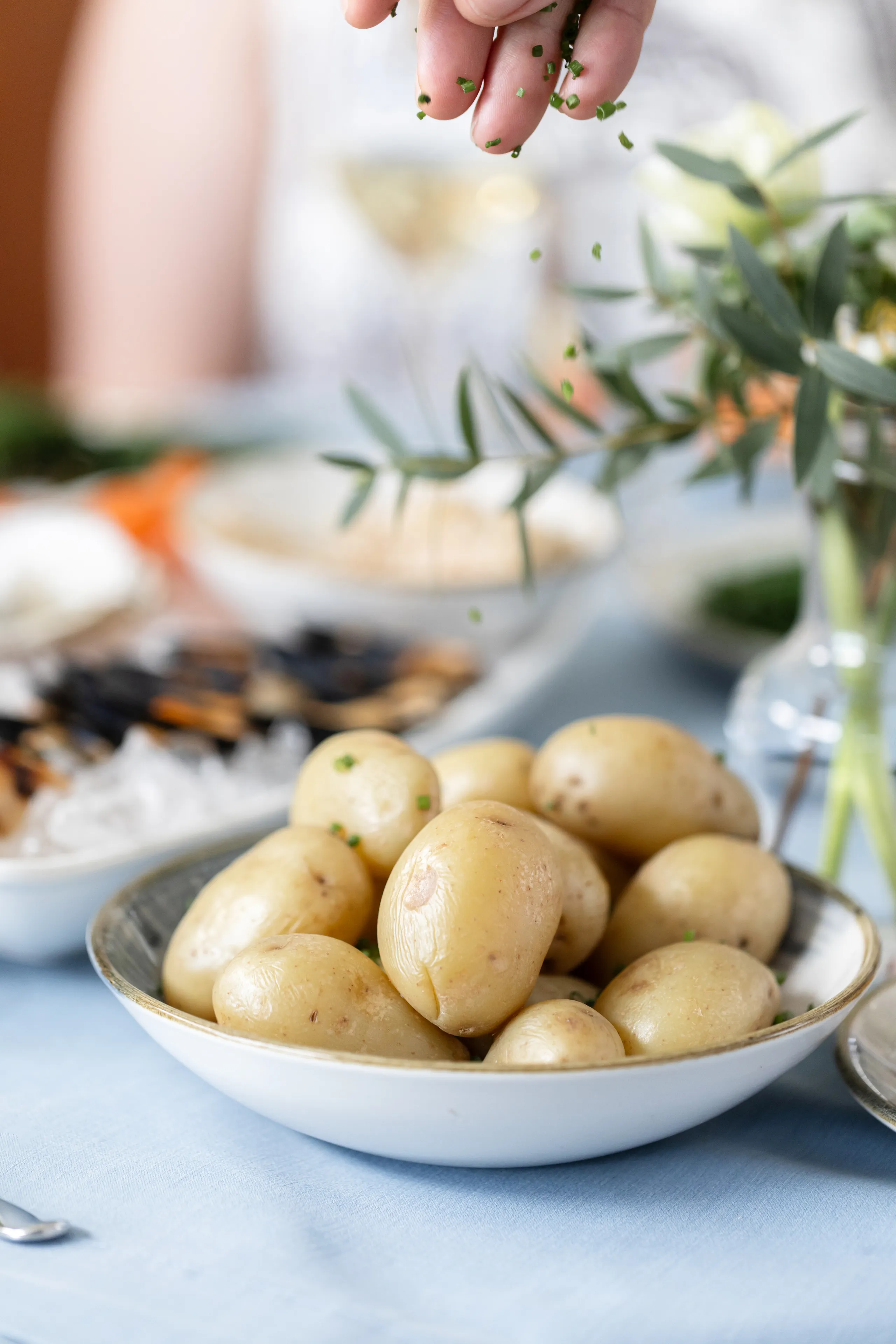 Hand sprinkling chopped chives over a bowl of boiled new potatoes on a table