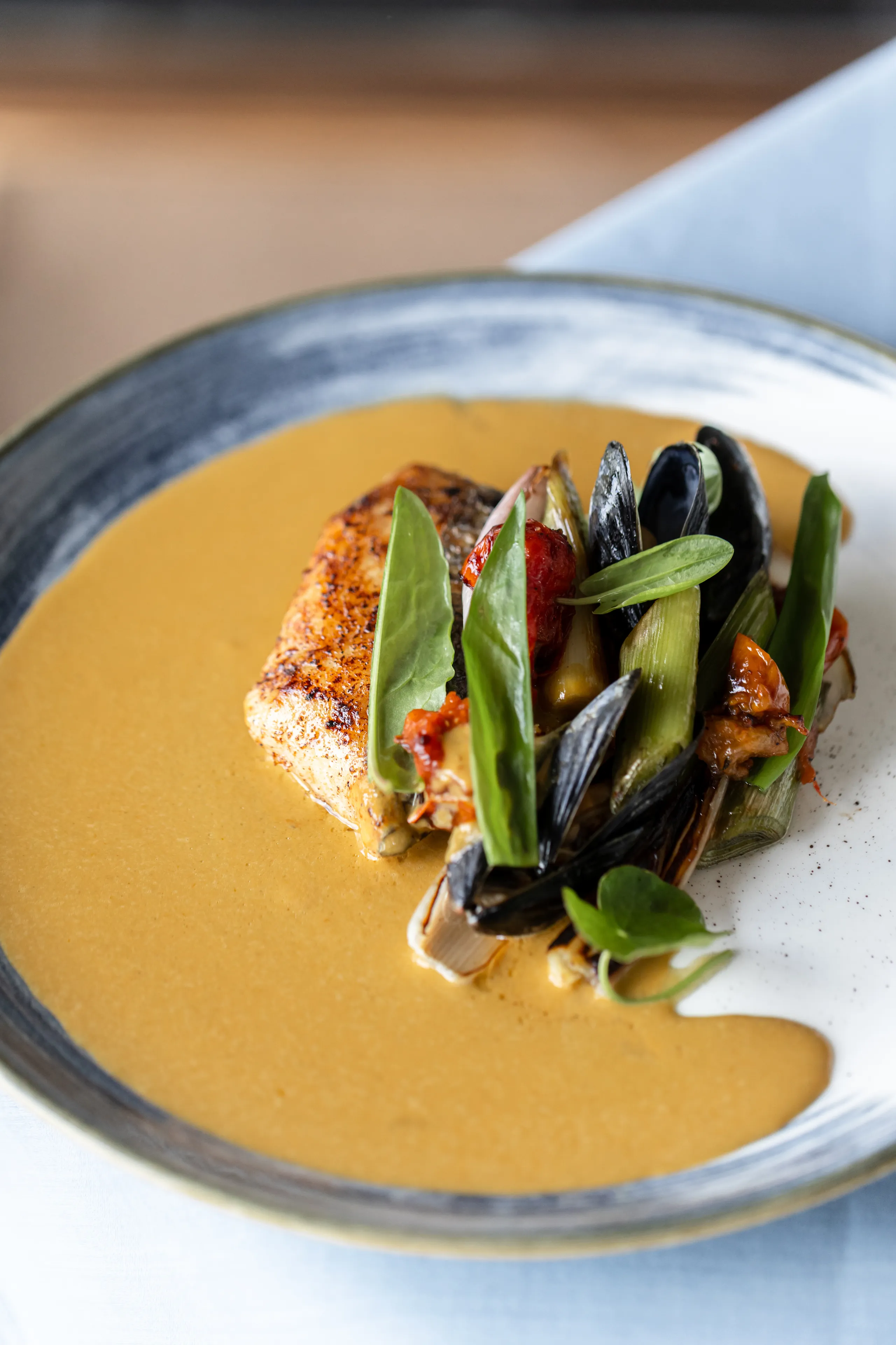 Pan-seared fish fillet served in a rich golden sauce with mussels, leeks, cherry tomatoes and fresh green leaves.