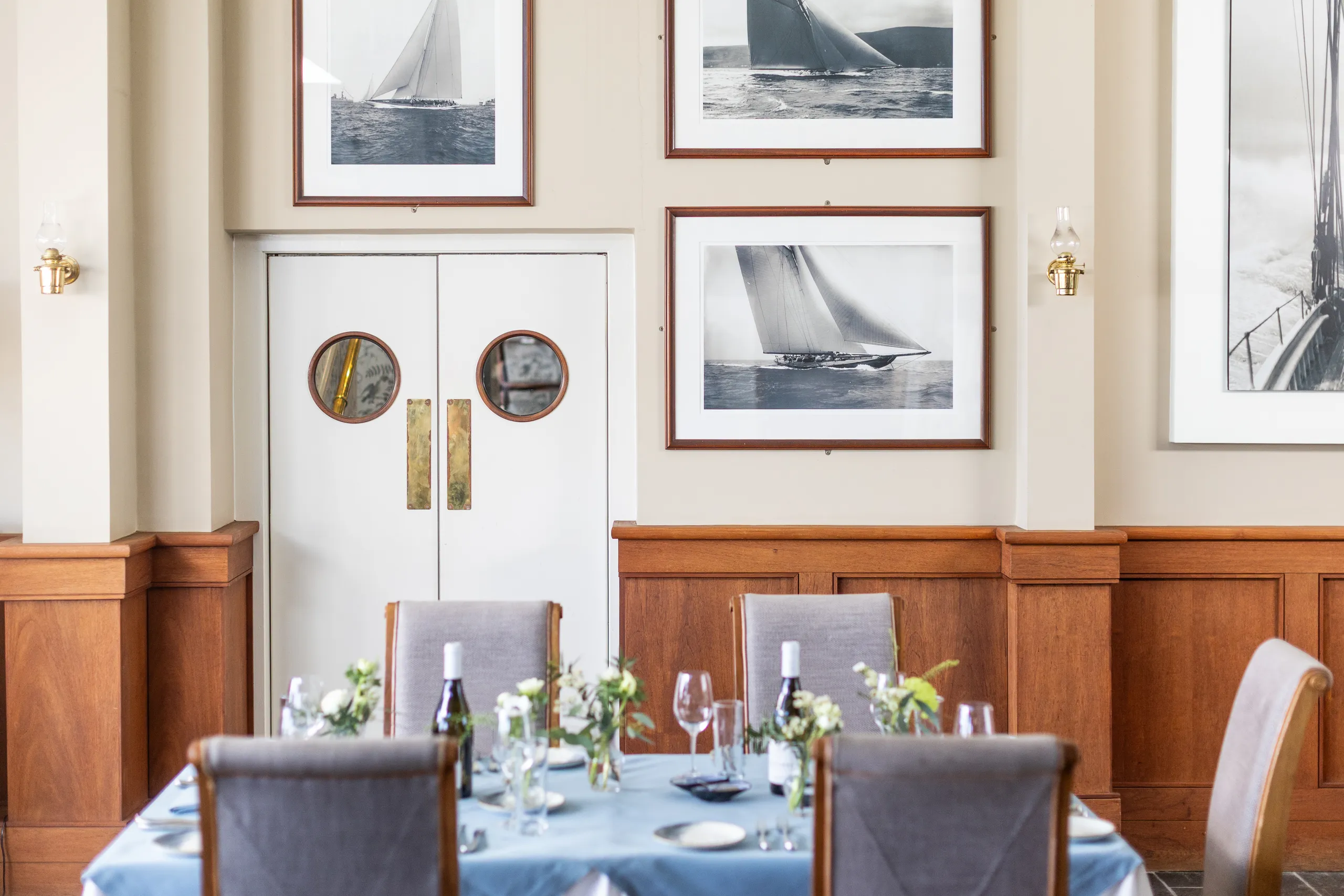 Elegant dining room with a laid table, framed sailing photographs and white double doors with round porthole windows.