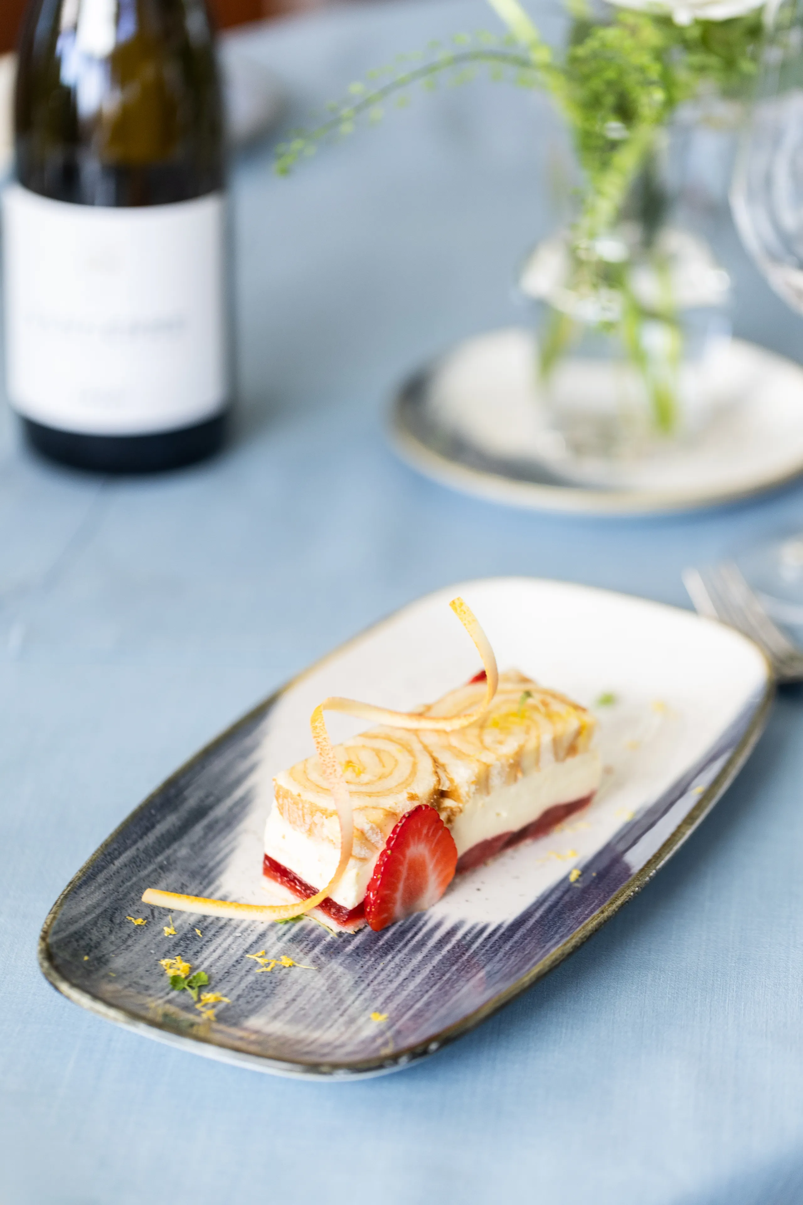 A plated layered dessert with toasted topping, strawberry garnish and citrus zest on a blue tablecloth, with a wine bottle and flowers softly blurred in the background.