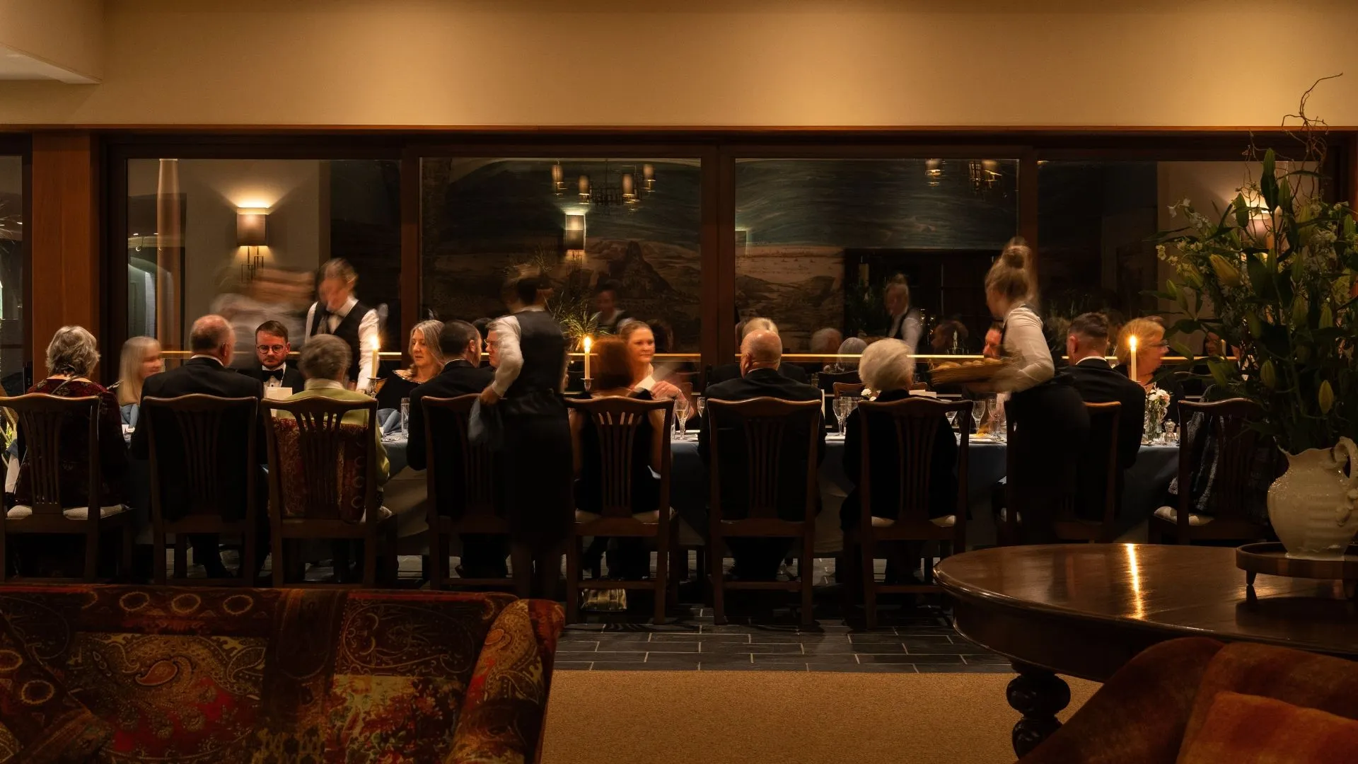 Guests seated at a long dining table in an elegant restaurant while waiters serve food in warm candlelight.