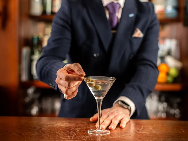 Bartender in a navy suit garnishing a martini with green olives at a wooden bar