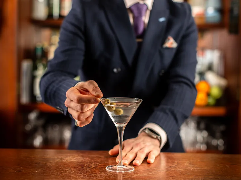 Bartender in a navy suit garnishing a martini with green olives at a wooden bar