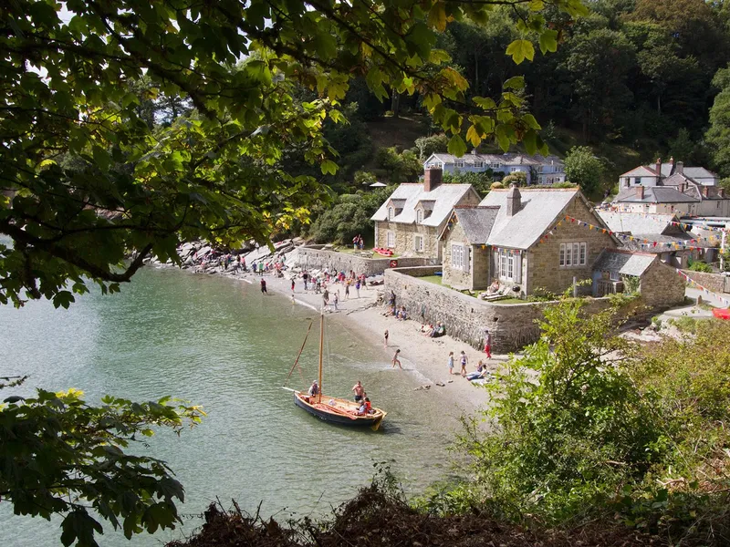Small sailing boat near a sandy beach and stone cottages in a sheltered Cornish cove, framed by leafy trees