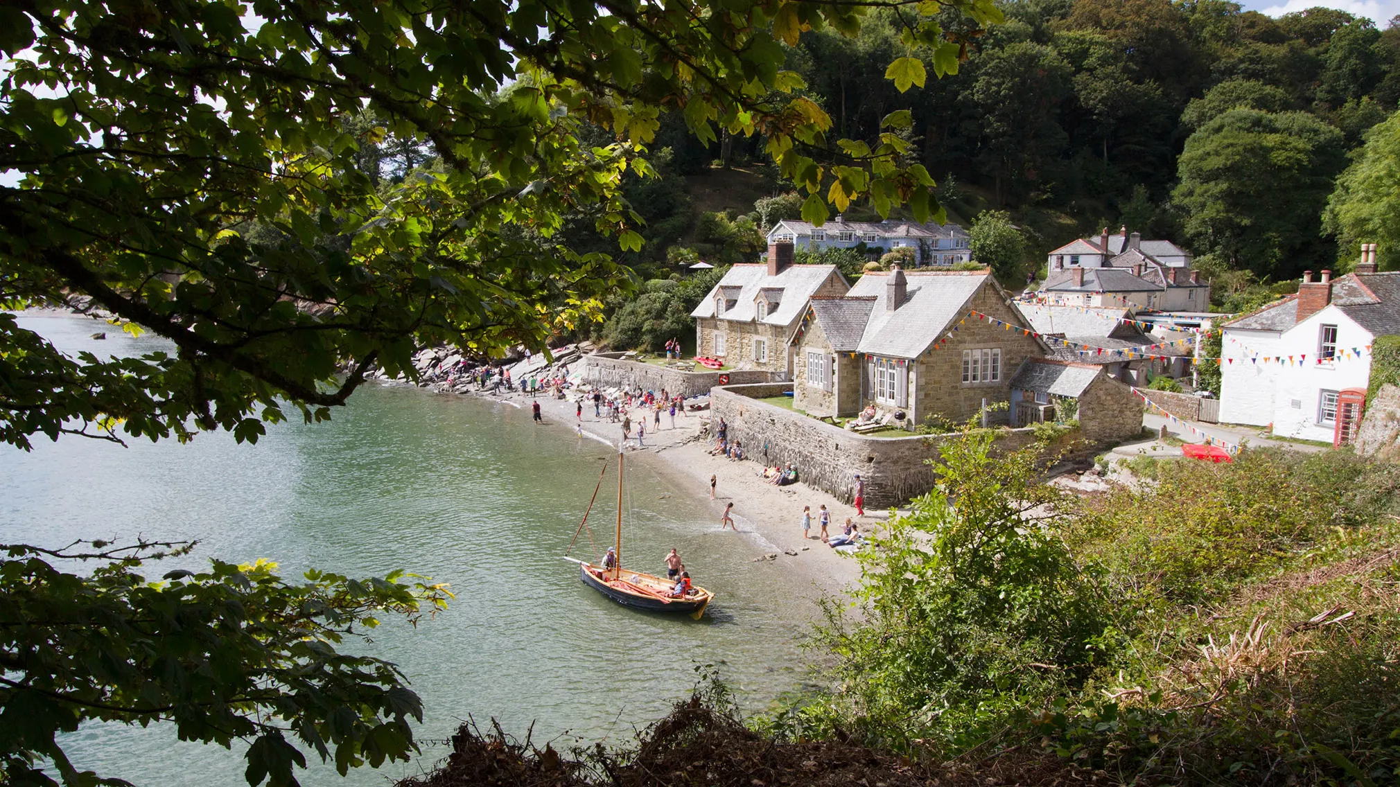 Small sailing boat near a sandy beach and stone cottages in a sheltered Cornish cove, framed by leafy trees