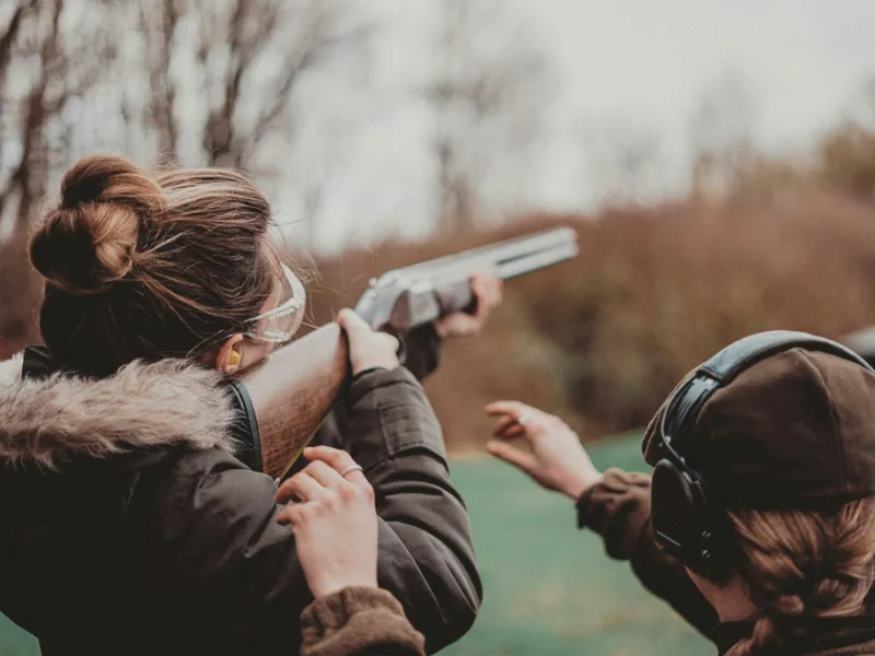 Person aiming a shotgun at an outdoor clay pigeon shooting range while another person stands beside them wearing ear defenders.