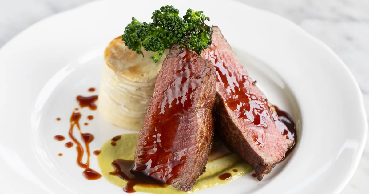 Slices of glazed pink duck breast served with a layered potato stack, broccolini and sauce on a white plate