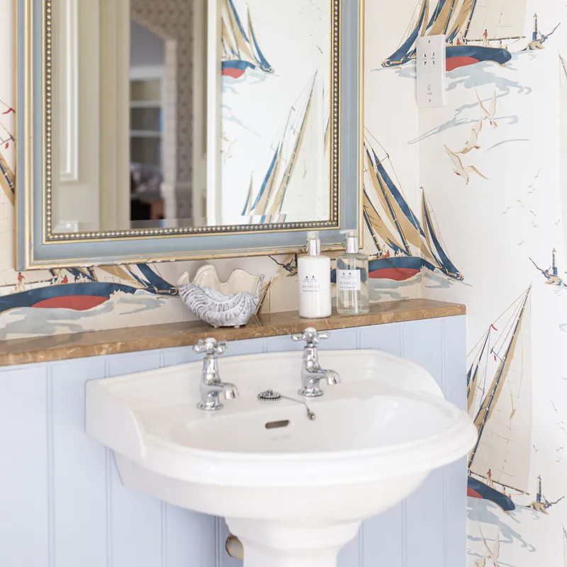 Pedestal basin beneath a framed mirror in a bathroom with nautical sailboat wallpaper