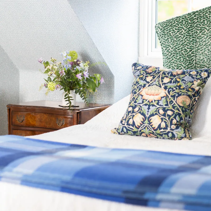 Elegant bedroom corner with patterned wallpaper, floral cushion, green upholstered headboard and wooden bedside table with fresh flowers