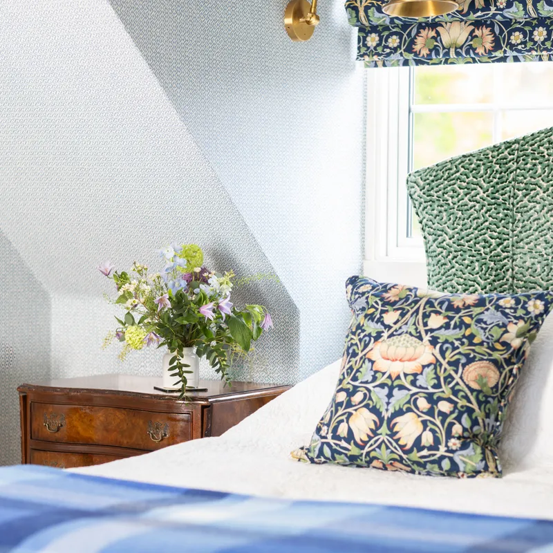 Elegant bedroom corner with patterned wallpaper, floral cushion, green upholstered headboard and wooden bedside table with fresh flowers