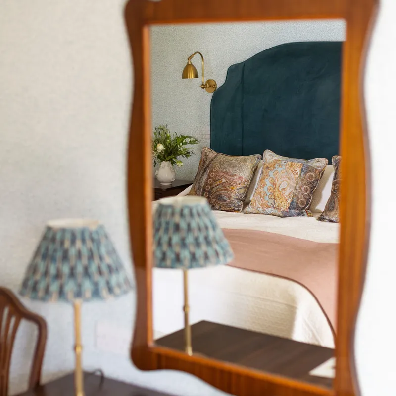 Mirror reflection of a stylish bedroom with a dark teal upholstered headboard, patterned cushions and brass wall light