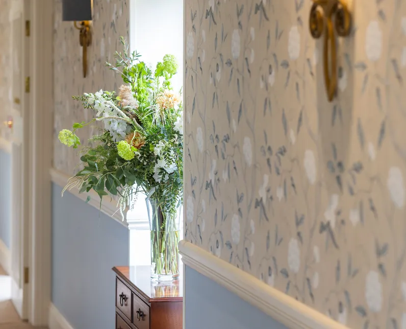 Elegant hallway with floral wallpaper, blue wainscoting, wall sconces and a vase of fresh flowers on a wooden chest of drawers