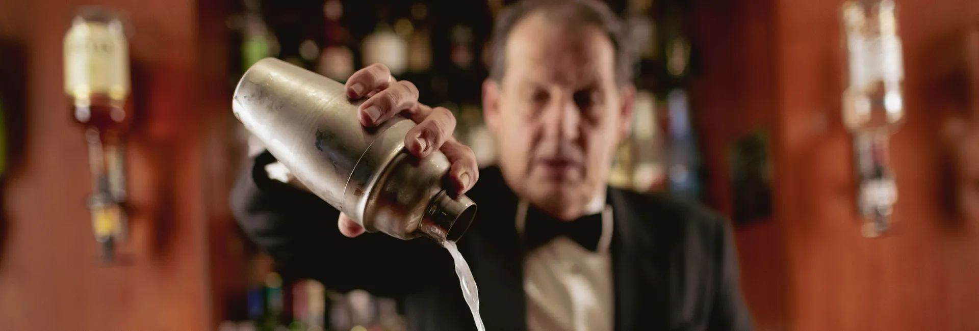 Bartender pouring a white cocktail from a shaker into a salt-rimmed martini glass with a lime wedge in a bar.