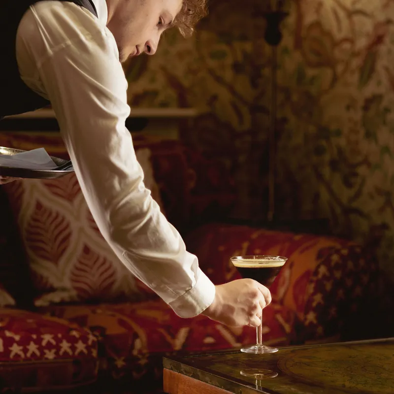 Waiter placing a dark espresso martini on a wooden table in a warmly lit lounge