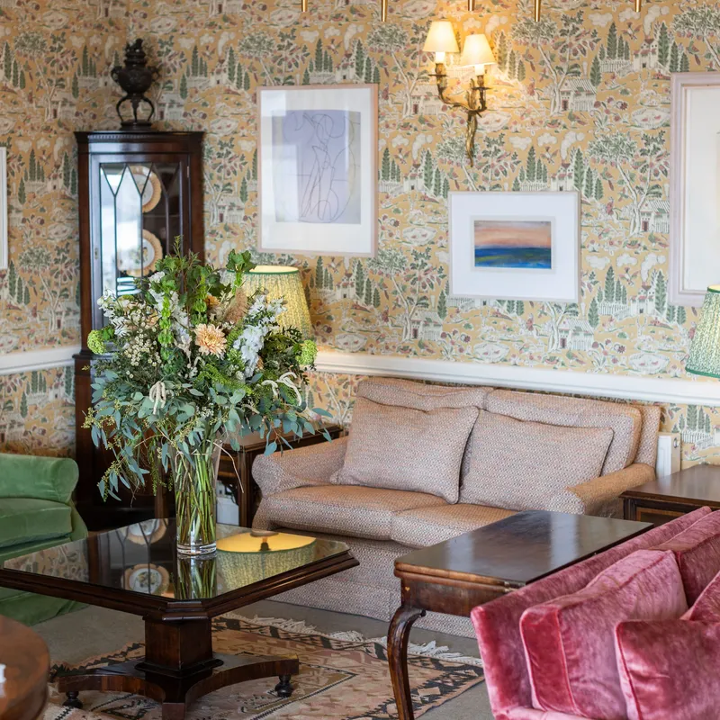 Elegant sitting room with patterned wallpaper, a brass chandelier, soft sofas, velvet armchairs and a large floral arrangement on a glass coffee table