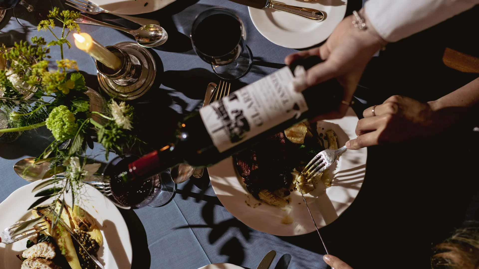 Overhead view of a candlelit dinner table with plates of food, red wine and a bottle being poured