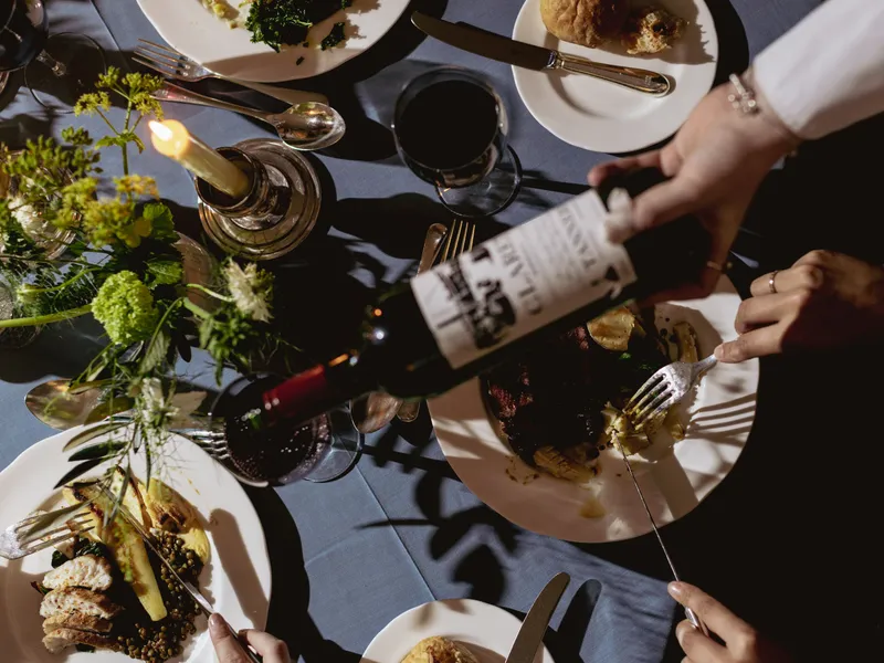 Overhead view of a candlelit dinner table with plates of food, red wine and a bottle being poured