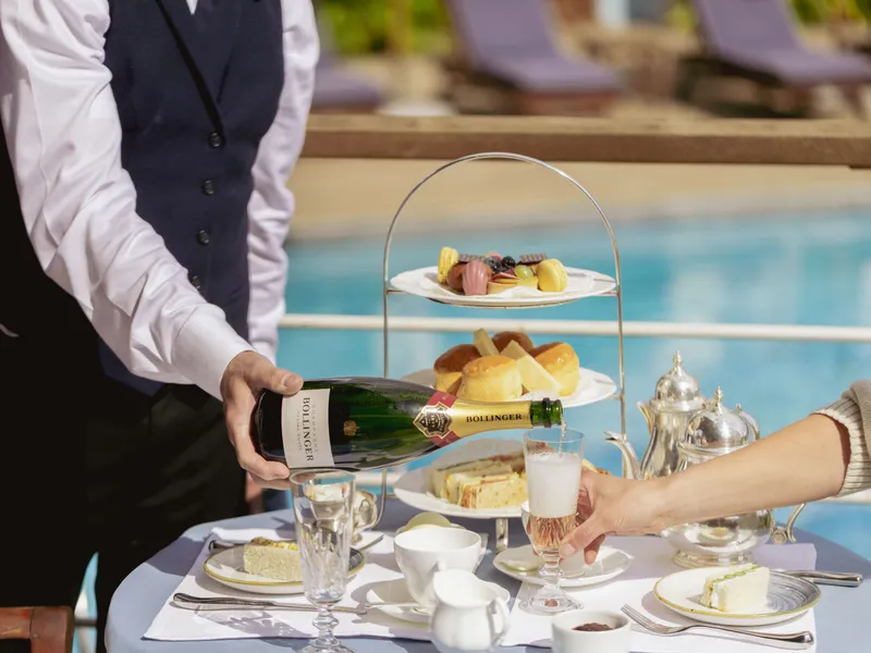 A waiter pours champagne into a glass beside a poolside afternoon tea with cakes, sandwiches and silver teapots.