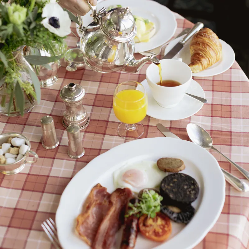 Silver teapot pouring tea at a breakfast table with a full cooked breakfast, croissant, orange juice and fresh flowers