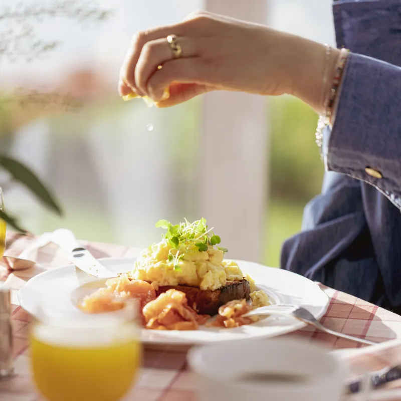 Hand squeezing lemon over scrambled eggs and smoked salmon on toast at a sunlit breakfast table.
