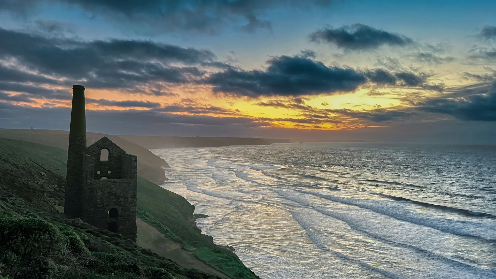 Ruined engine house and chimney on a cliff above a windswept beach at sunset