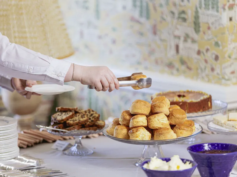 Person serving a scone with tongs from a tiered plate at an afternoon tea buffet