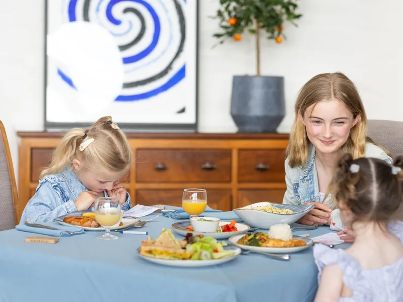 Three children sitting at a dining table with plates of food and glasses of juice in a bright home dining room.