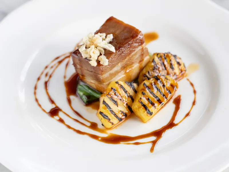 Plated pork belly with grilled pineapple, green vegetables and a rich sauce drizzle