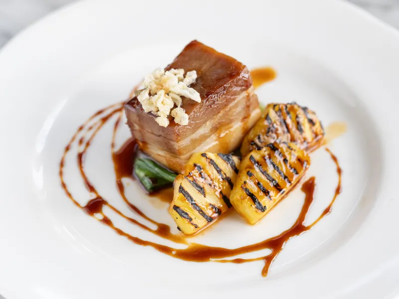 Plated pork belly with grilled pineapple, green vegetables and a rich sauce drizzle