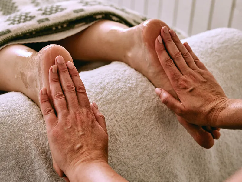 Close-up of a therapist applying massage oil to a client's feet on a towel-covered treatment bed.