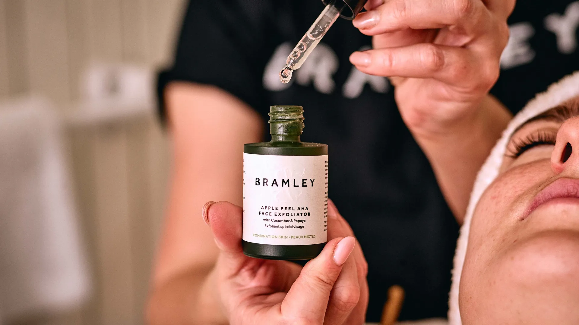 Beauty therapist holding a Bramley apple peel face exfoliator bottle and glass dropper beside a client's face.