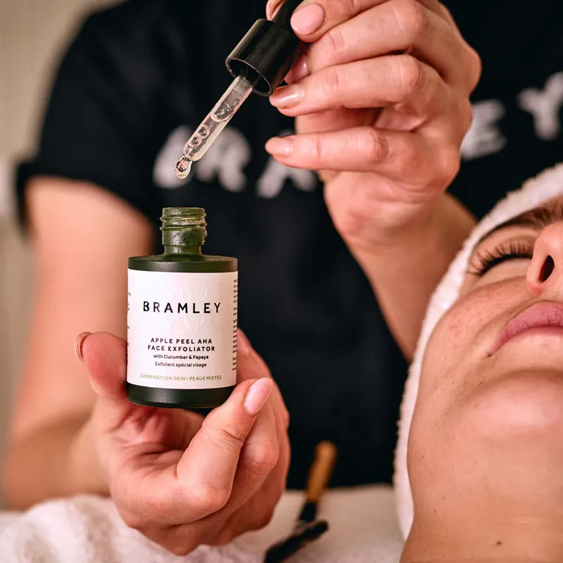 Beauty therapist holding a Bramley apple peel face exfoliator bottle and glass dropper beside a client's face.