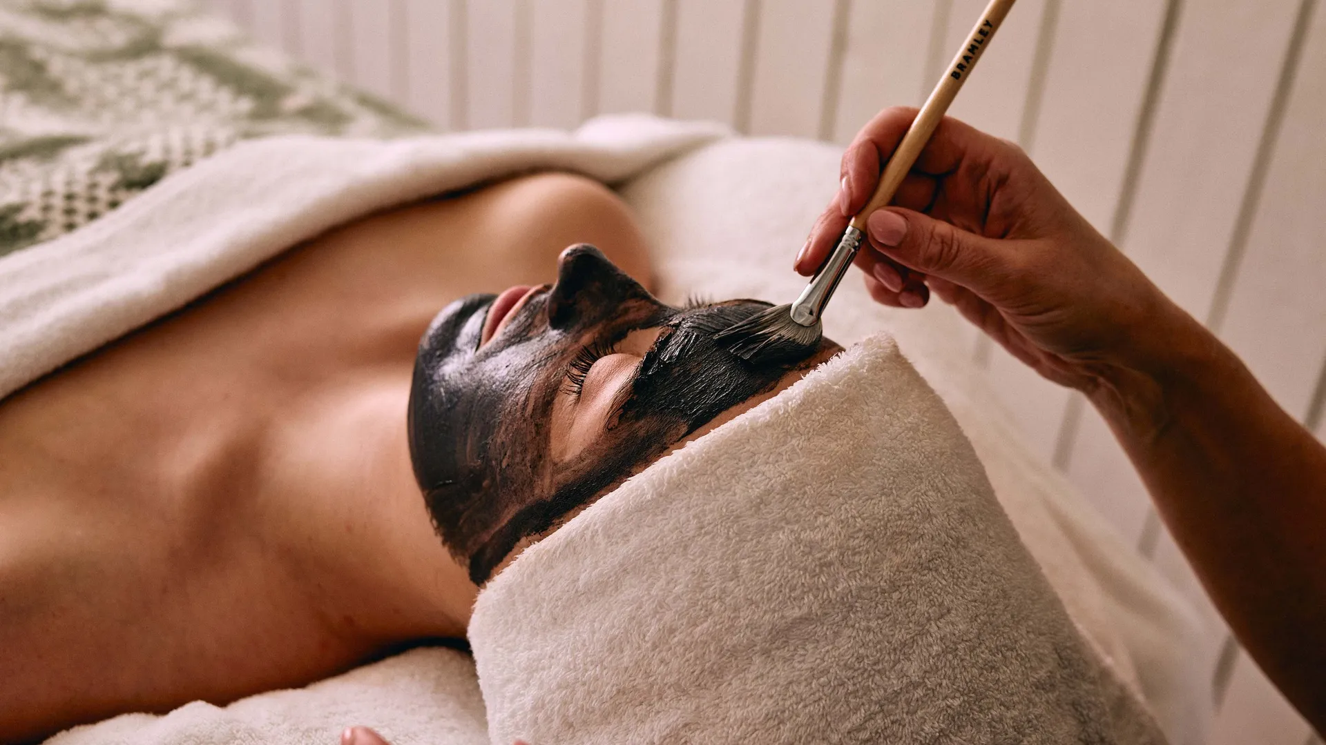 Person receiving a dark facial mask applied with a brush during a spa treatment.