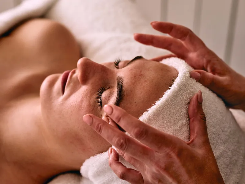 Woman receiving a relaxing facial massage while lying on a treatment bed with a towel headband.