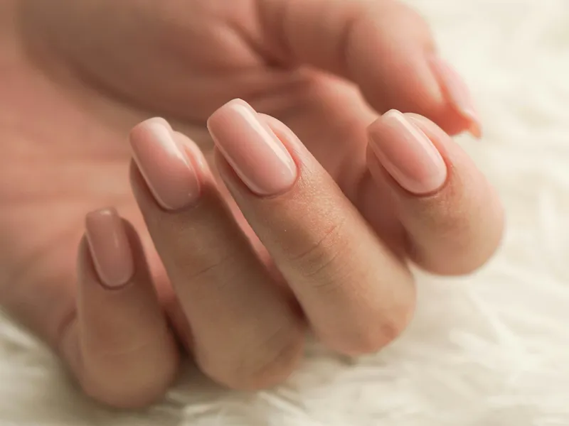 Close-up of hands with glossy nude pink manicure resting on a soft white surface beside a blurred mirror.