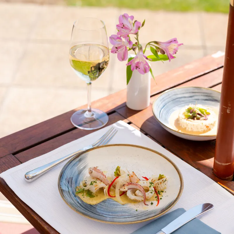 Outdoor restaurant table set with squid dish, white wine and pink flowers in sunlight