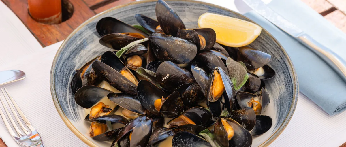 Bowl of steamed mussels with lemon beside a mint cocktail on an outdoor table
