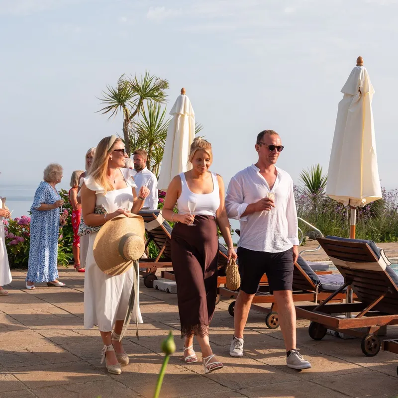 Guests holding drinks at a sunny poolside gathering overlooking the sea.