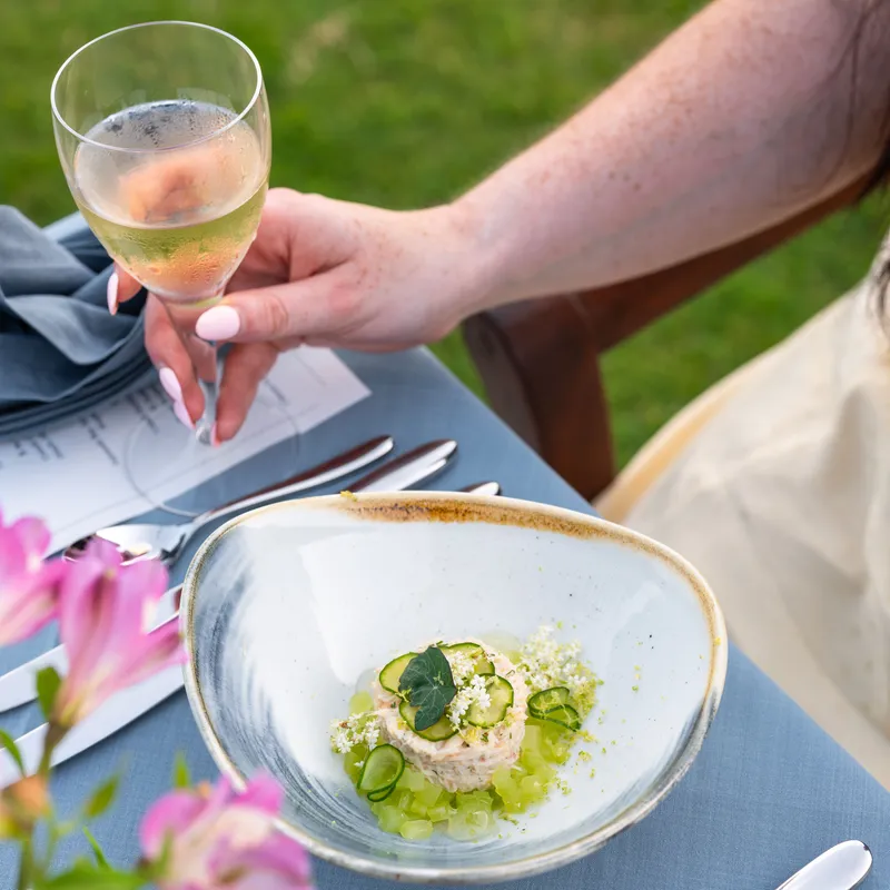 A woman holding a glass of sparkling wine beside a plated cucumber-topped starter at an outdoor table.