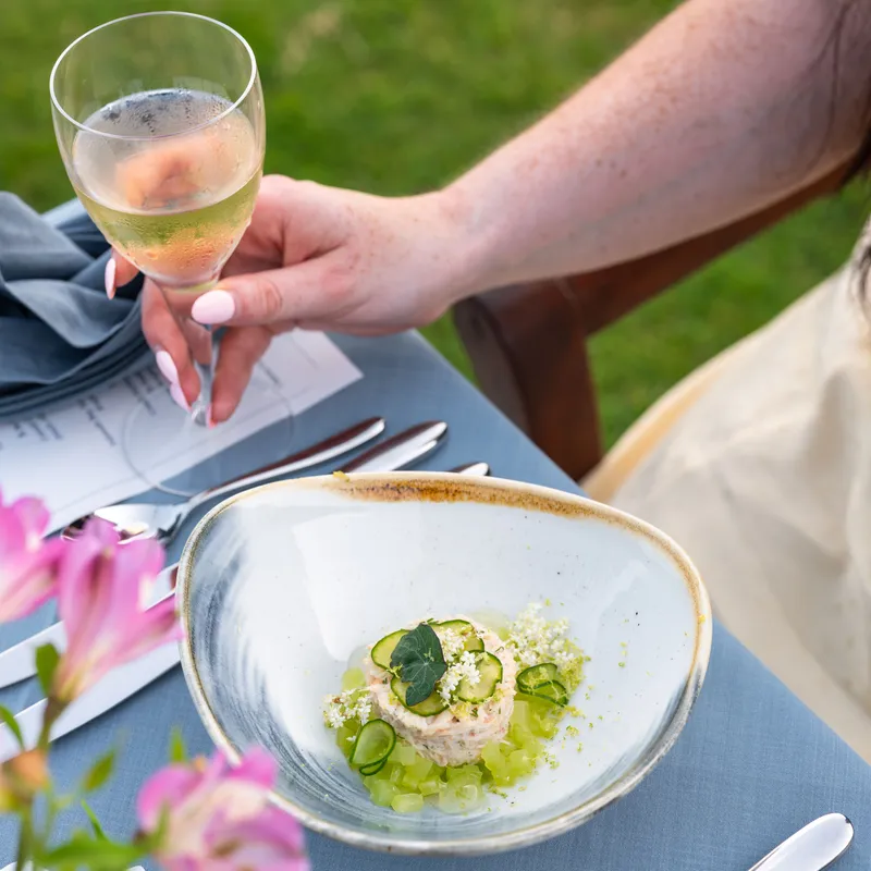 A woman holding a glass of sparkling wine beside a plated cucumber-topped starter at an outdoor table.