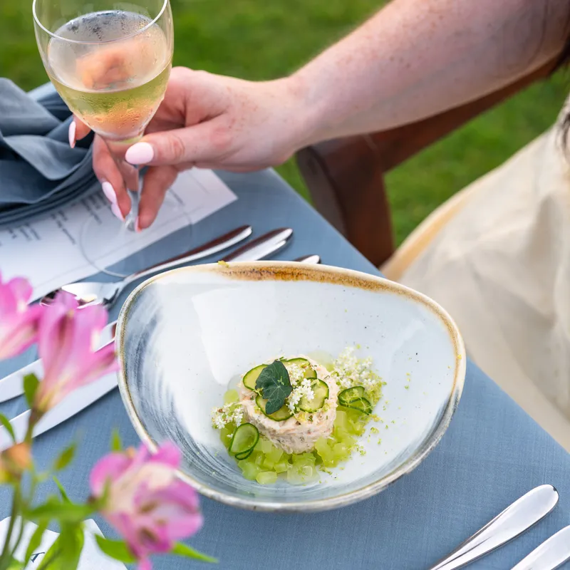 A woman holding a glass of sparkling wine beside a plated cucumber-topped starter at an outdoor table.