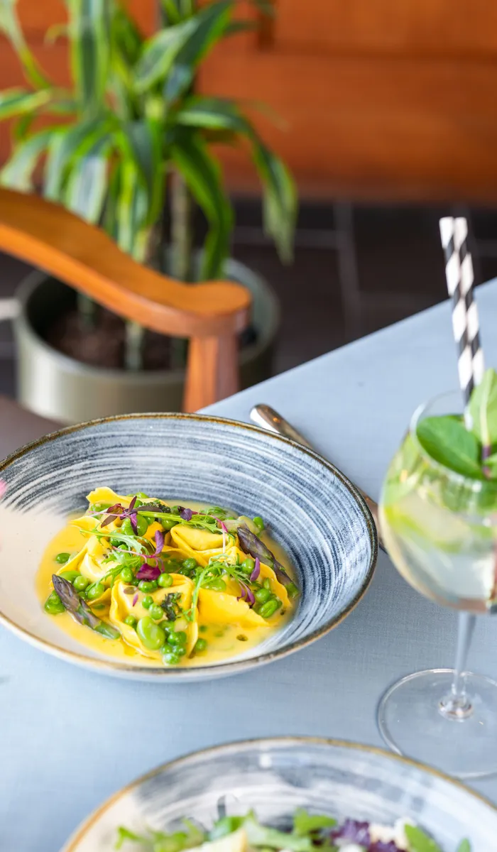 Bowl of tortellini with peas, asparagus and microgreens on a restaurant table beside a fresh drink.