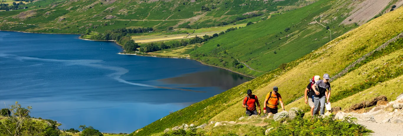 Four hikers climbing a rocky mountain path above a deep blue lake and green valley.