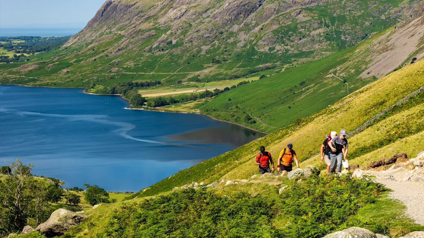 Four hikers climbing a rocky mountain path above a deep blue lake and green valley.