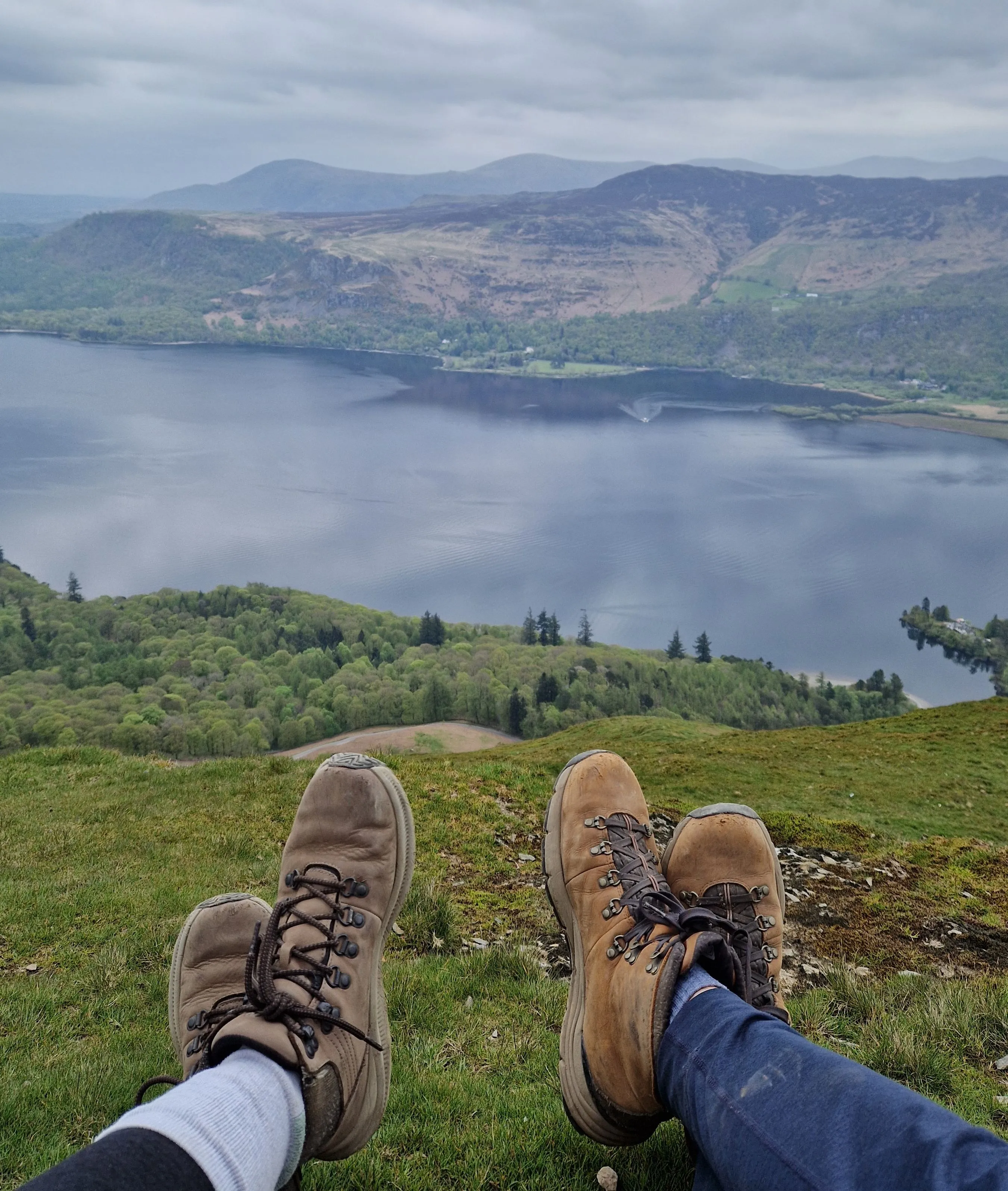 Two hikers in boots resting on a grassy hill overlooking a lake and misty mountains beneath cloudy skies.