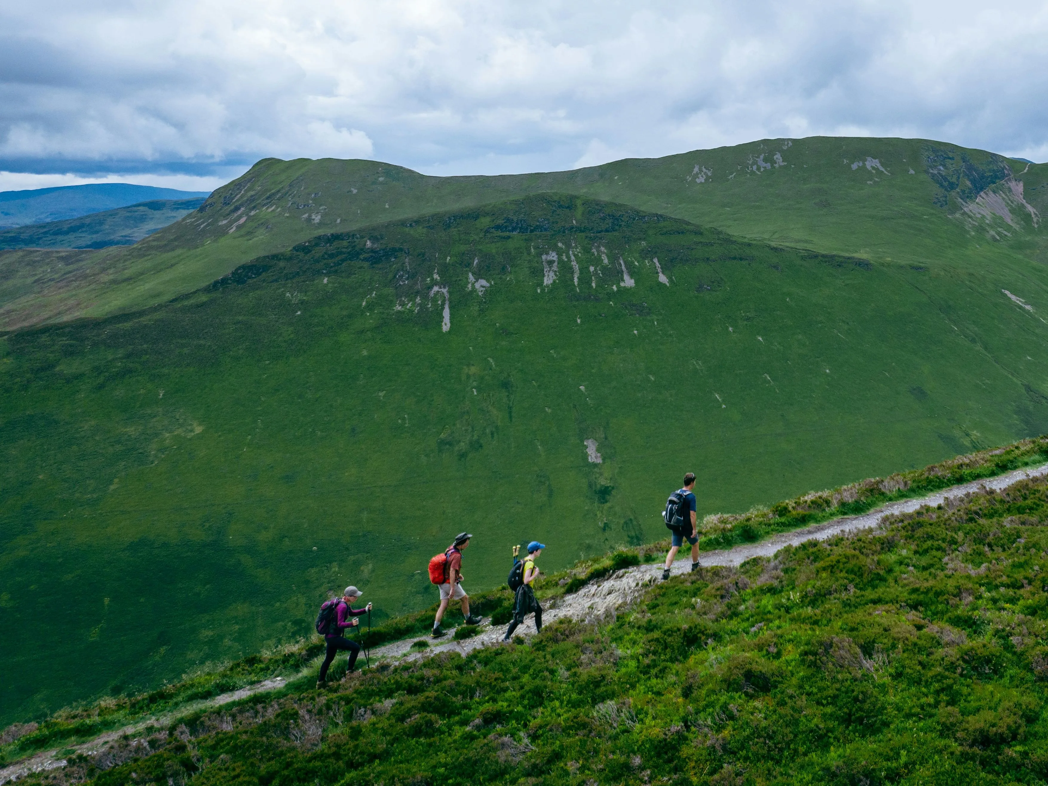 Four hikers walking along a narrow trail across a green mountain hillside beneath a cloudy sky.